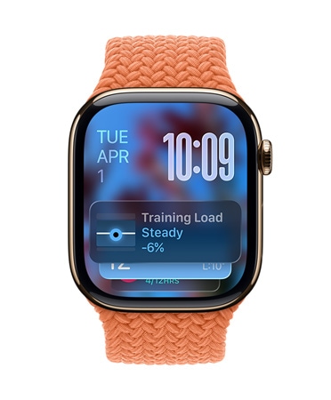 R??ově zlaté Apple?Watch s?mandarinkovym pletenym ?emínkem, v?levém horním rohu ciferníku se zobrazuje datum, vpravo naho?e ?as psany velkymi ?íslicemi, dole widget Tréninková zátě?, to v?echno v?pr?svitnémm Liquid Glass designu