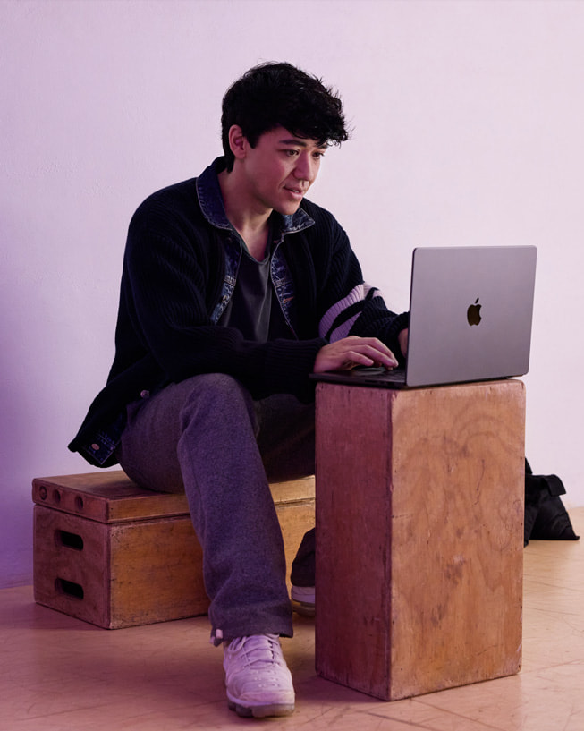 Kyle Hanagami sitzt in einem Studio auf einer Kiste und tippt auf einem MacBook Pro, das auf einer weiteren Kiste steht.