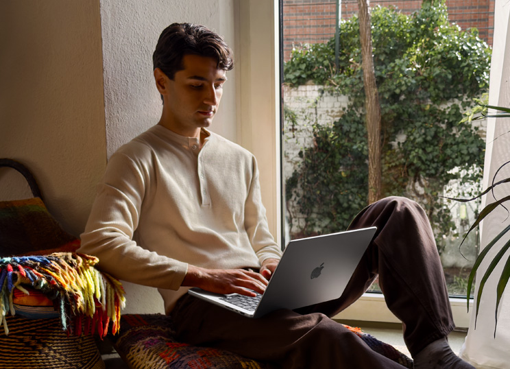 Une personne assise par terre à c?té d’une fenêtre utilisant un MacBook.