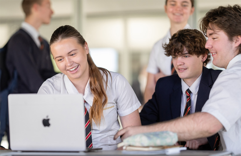 Eine Gruppe von Schüler:innen in Schuluniformen versammelt sich um ein MacBook, l?chelt und arbeitet zusammen.