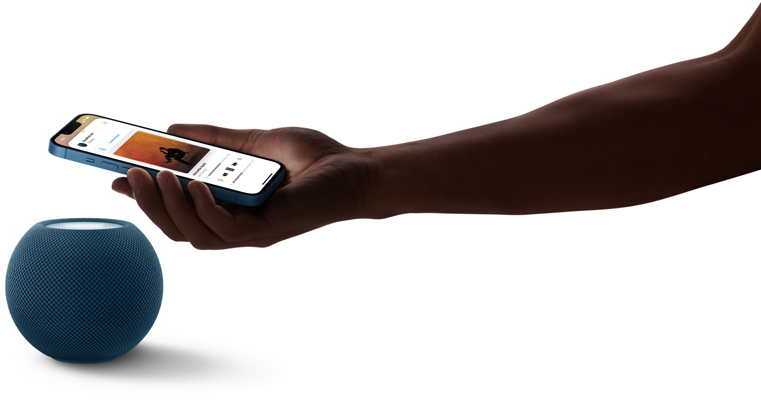 Der HomePod mini in Blau und eine Hand, die ein iPhone über den HomePod mini h?lt. Auf dem iPhone Display wird Musik abgespielt.