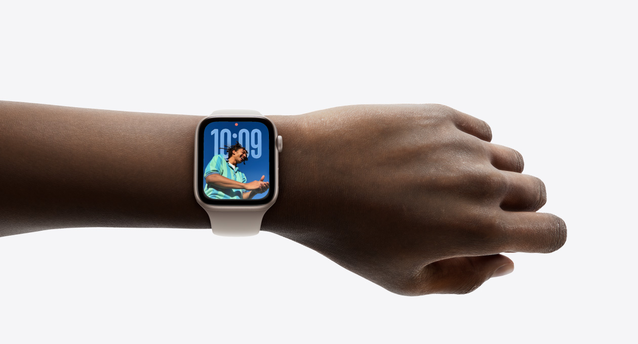 Pohled zblízka na st?íbrné Apple?Watch s?gumovym ?emínkem nasazené na zápěstí, je vidět aplikace Hodiny v?novém Liquid Glass designu